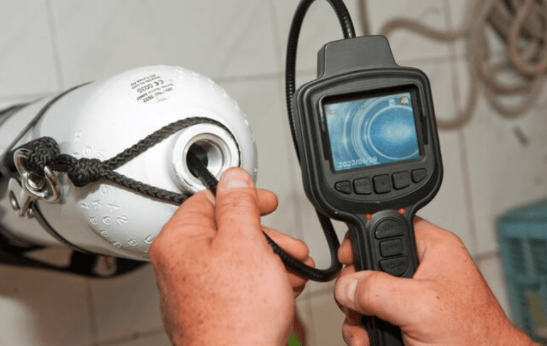 A person uses a handheld inspection camera to examine the interior of a white metal cylinder, possibly a gas or diving cylinder, in a tiled room. The camera screen displays an image from inside the cylinder.