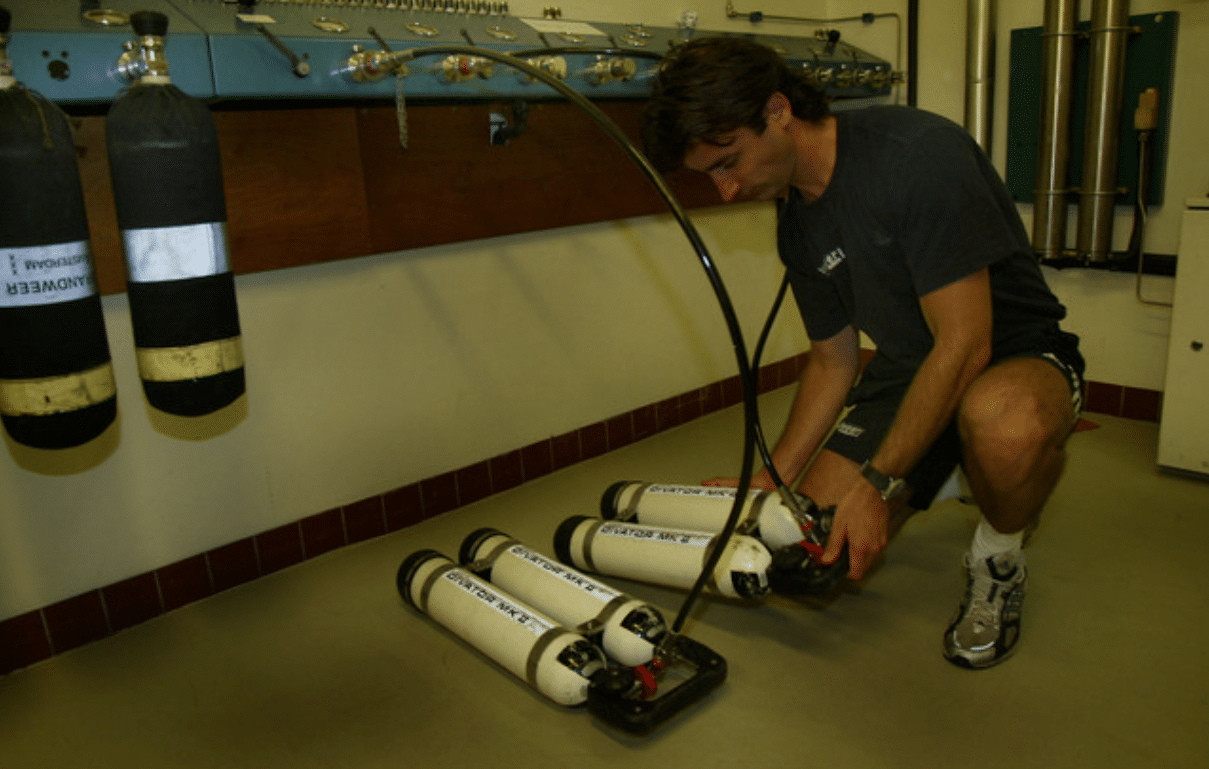 Screenshot 2026-04-06 140756 - N9BO℠ | Global Underwater Services Ltd A person kneels on the floor, connecting air cylinders with hoses in a lab setting; more gas cylinders hang on the wall in the background.