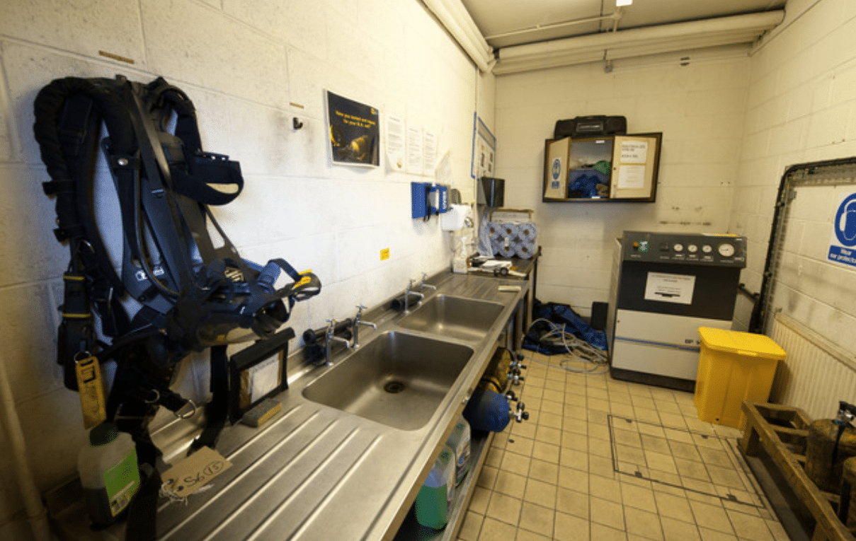 Screenshot 2026-04-06 141522 - N9BO℠ | Global Underwater Services Ltd A small, well-lit room with stainless steel sinks, scuba diving gear hanging on the left wall, cleaning supplies, a yellow bin, and equipment for servicing scuba cylinders on the right. The floor is tiled.