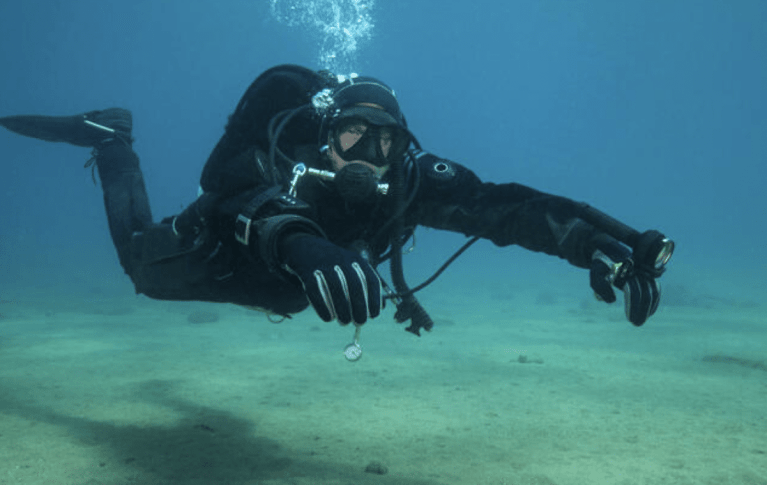A scuba diver in a black wetsuit and mask swims underwater near the sandy sea floor, holding a compass and torch while bubbles rise above their head.