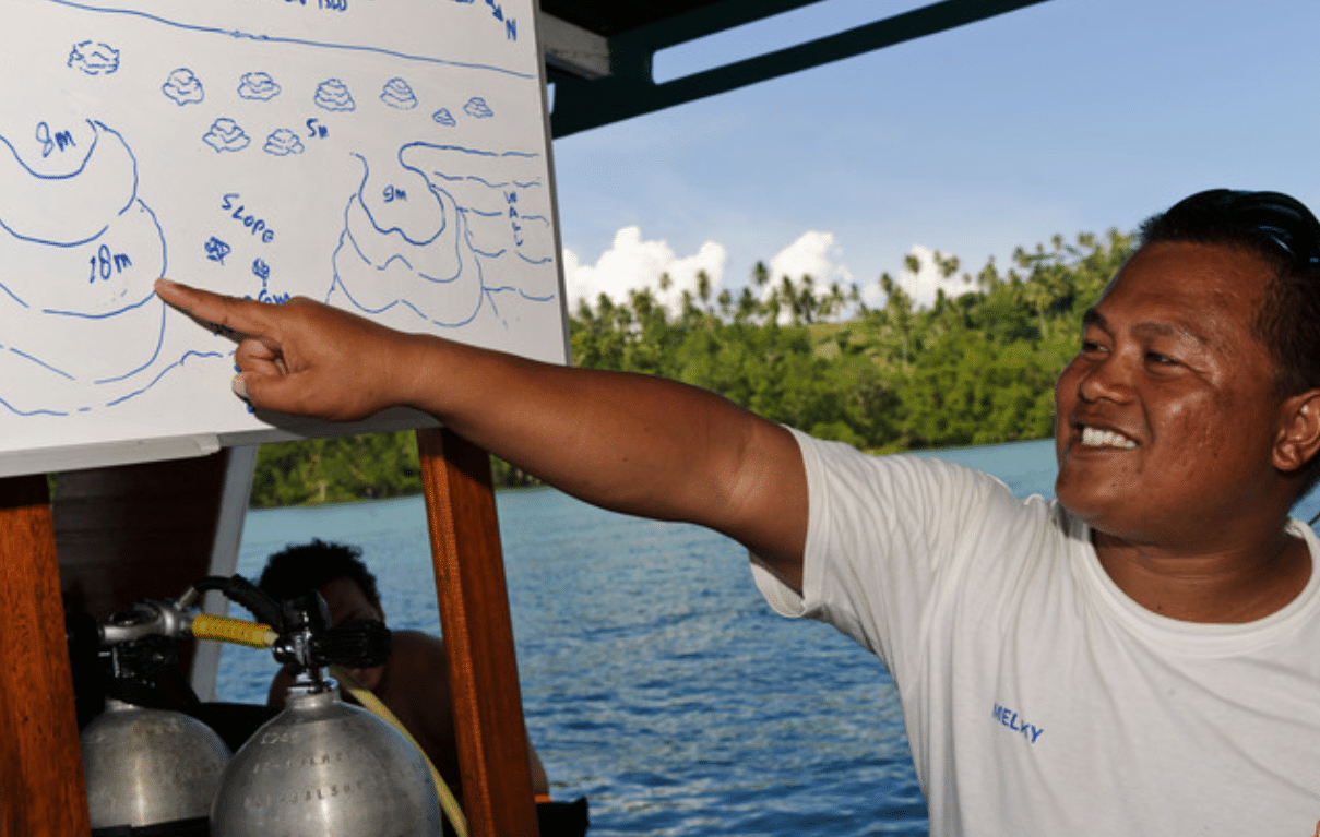A smiling man in a white shirt points at a whiteboard with hand-drawn underwater topography, likely explaining a dive site, with scuba cylinders and ocean scenery in the background.