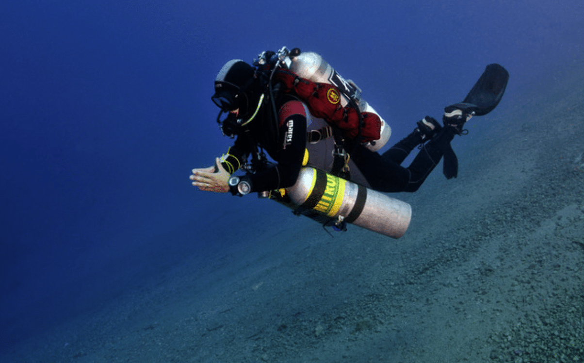 A scuba diver in a black wetsuit and full kit, including multiple cylinders, swims underwater above a sandy sea floor in clear blue water.