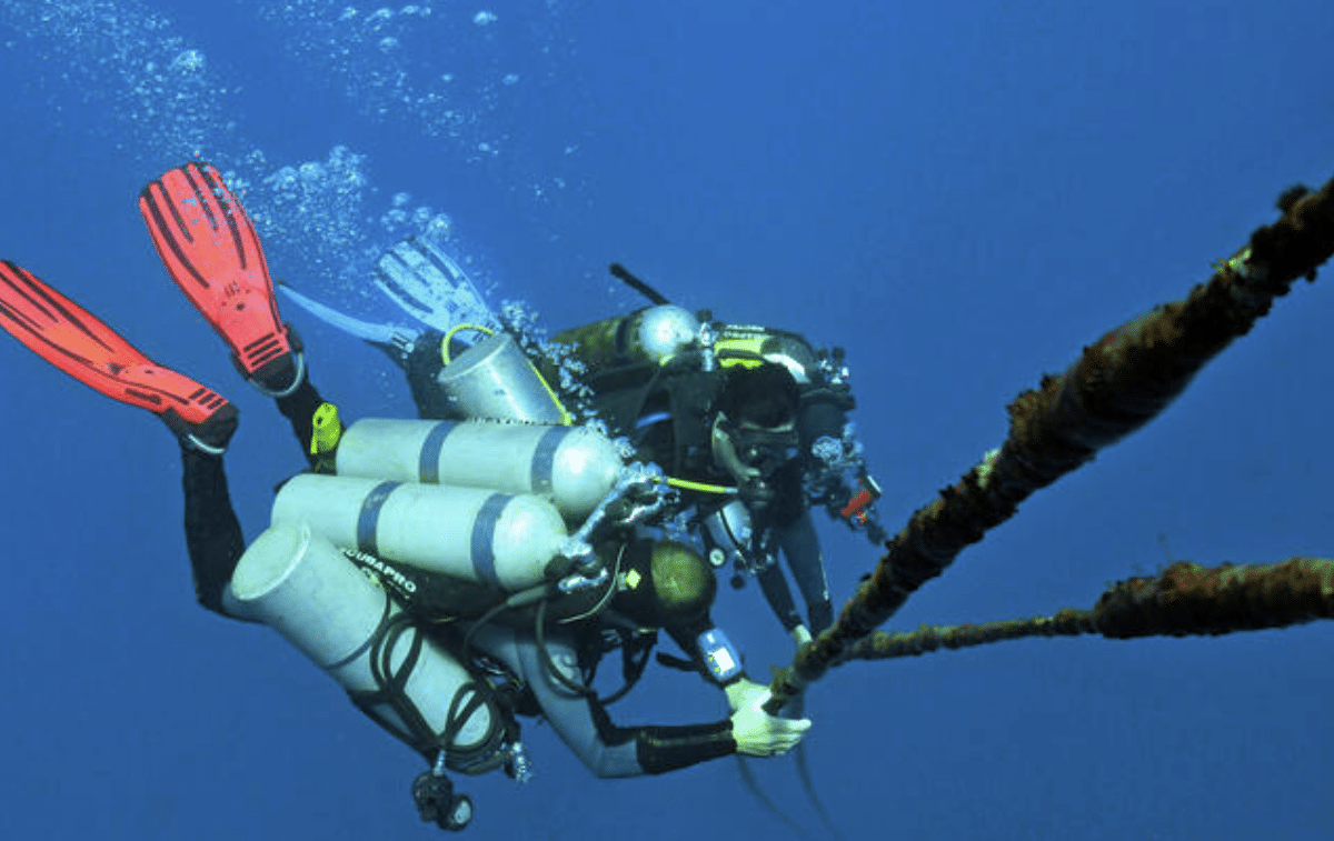Screenshot 2026-04-06 144201 - N9BO℠ | Global Underwater Services Ltd Two scuba divers equipped with multiple oxygen cylinders hold onto a thick, rusted underwater cable suspended in deep blue water, surrounded by bubbles.