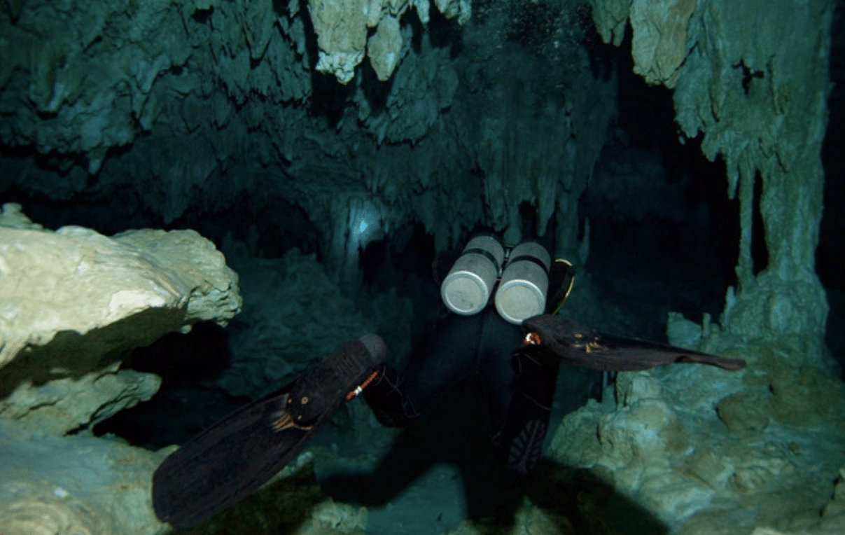 Screenshot 2026-04-06 154022 - N9BO℠ | Global Underwater Services Ltd A scuba diver with oxygen cylinders and fins swims through an underwater cave surrounded by jagged rock formations and stalactites.