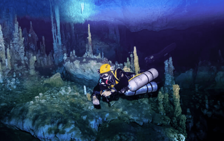 A scuba diver wearing a yellow helmet and carrying multiple cylinders explores an underwater cave filled with stalagmites and rocky formations, illuminated by blue and purple light.