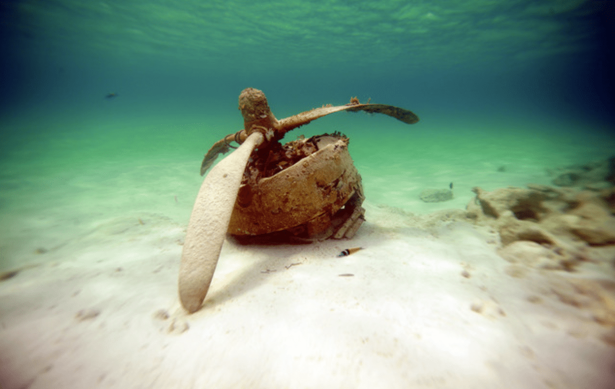 Screenshot 2026-04-06 155014 - N9BO℠ | Global Underwater Services Ltd An underwater photo of a rusted aeroplane propeller and engine resting on the sandy ocean floor, surrounded by clear blue-green water and sparse marine life.