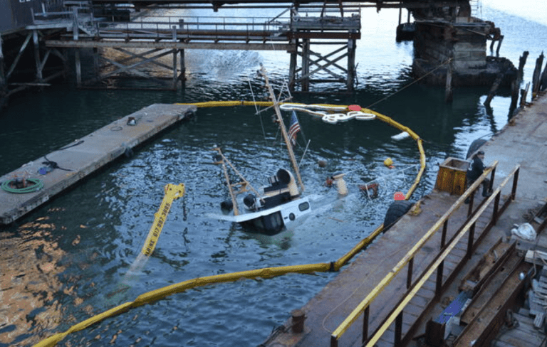 A partially submerged boat tilts in the water near a quay, surrounded by floating barriers. Part of its cabin and mast remain above water; an American flag is visible among debris and nearby construction.
