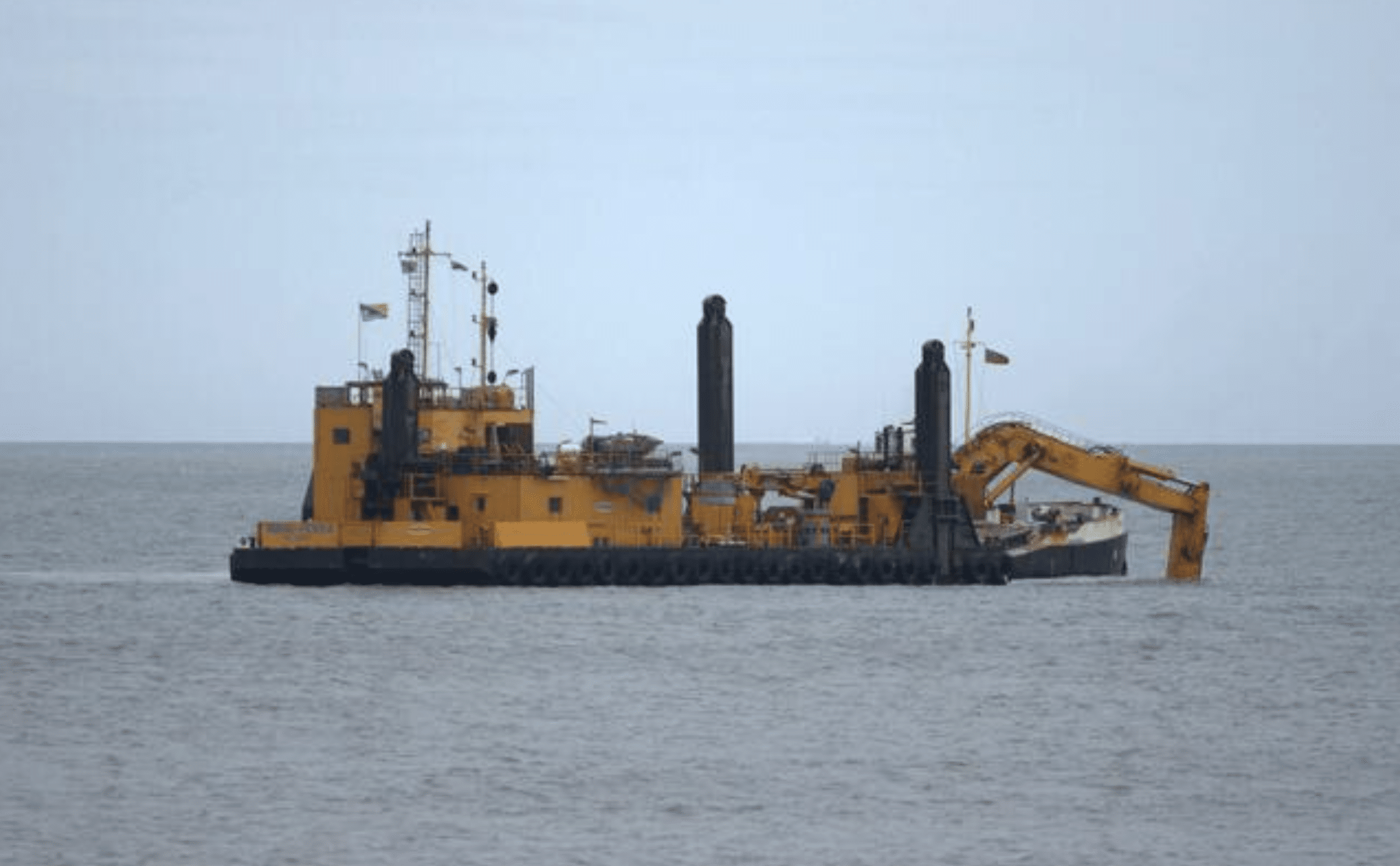 Screenshot 2026-04-06 at 190417 - N9BO℠ | Global Underwater Services Ltd A large yellow dredging vessel with machinery and cranes is floating on calm grey water under an overcast sky.