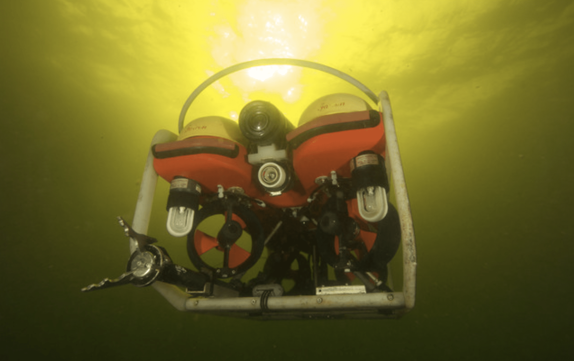 A remotely operated underwater vehicle (ROV) with red flotation devices is submerged underwater, illuminated by bright yellow-green light filtering down from the surface above.