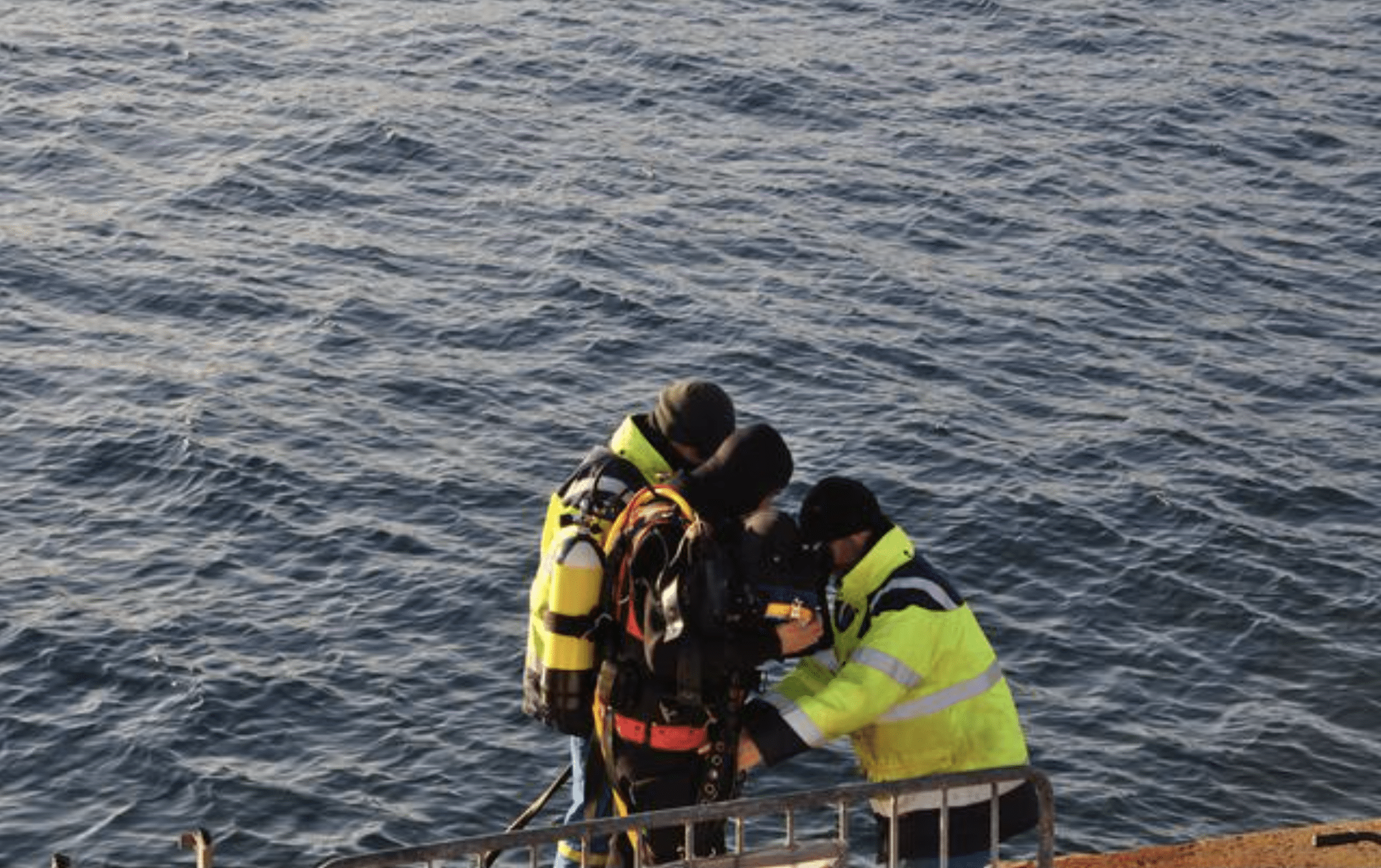 Two people in yellow high-visibility jackets help a diver prepare near the edge of a body of water, with safety equipment and railing visible in the foreground.