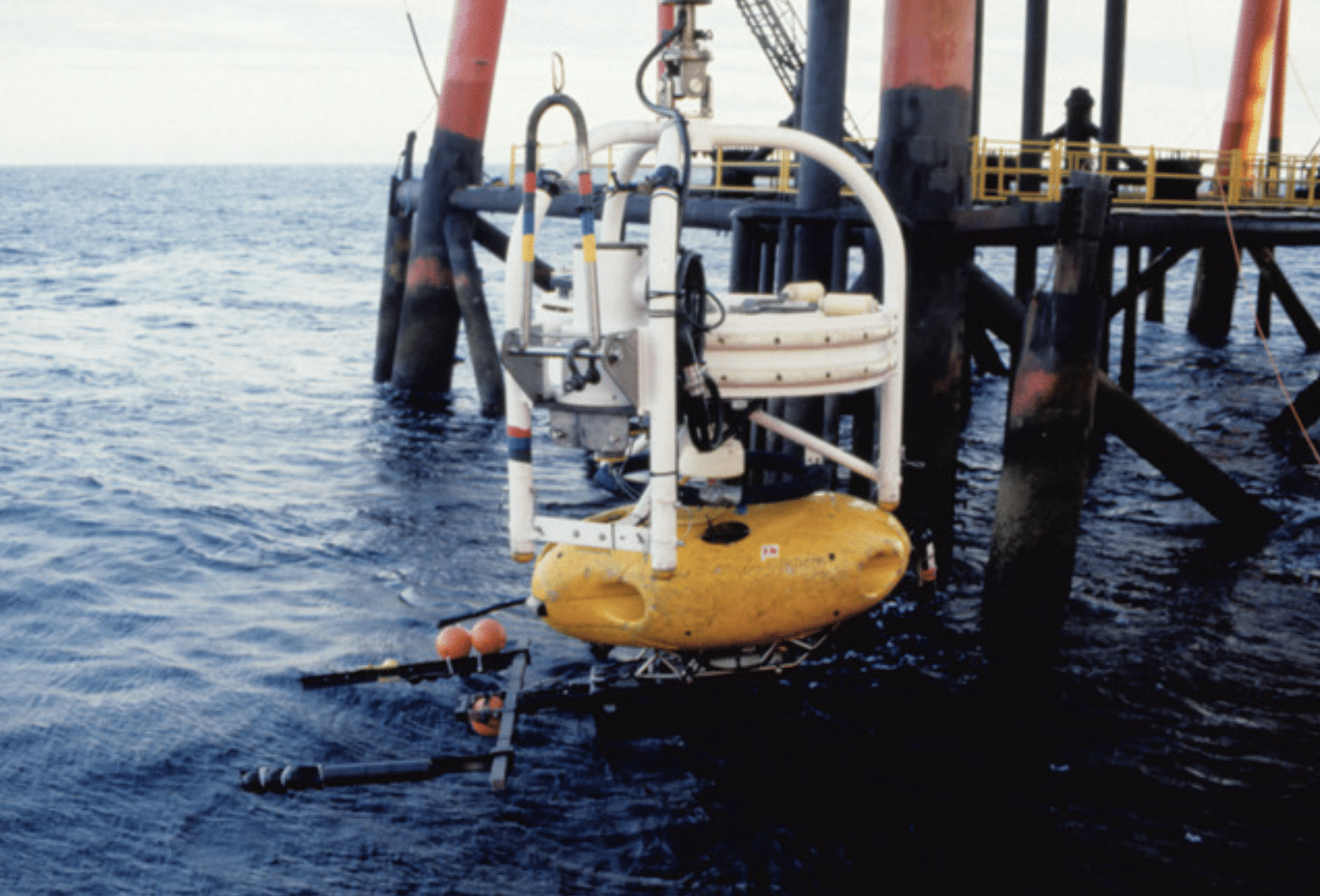 A yellow underwater remotely operated vehicle (ROV) is being lowered into the sea near a large structure with red and black support pillars. The water is calm and the structure appears industrial.