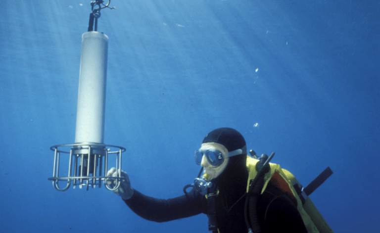 A scuba diver underwater wearing a wetsuit and mask holds a cylindrical scientific instrument attached to a metal frame, with beams of sunlight filtering through the blue water above.