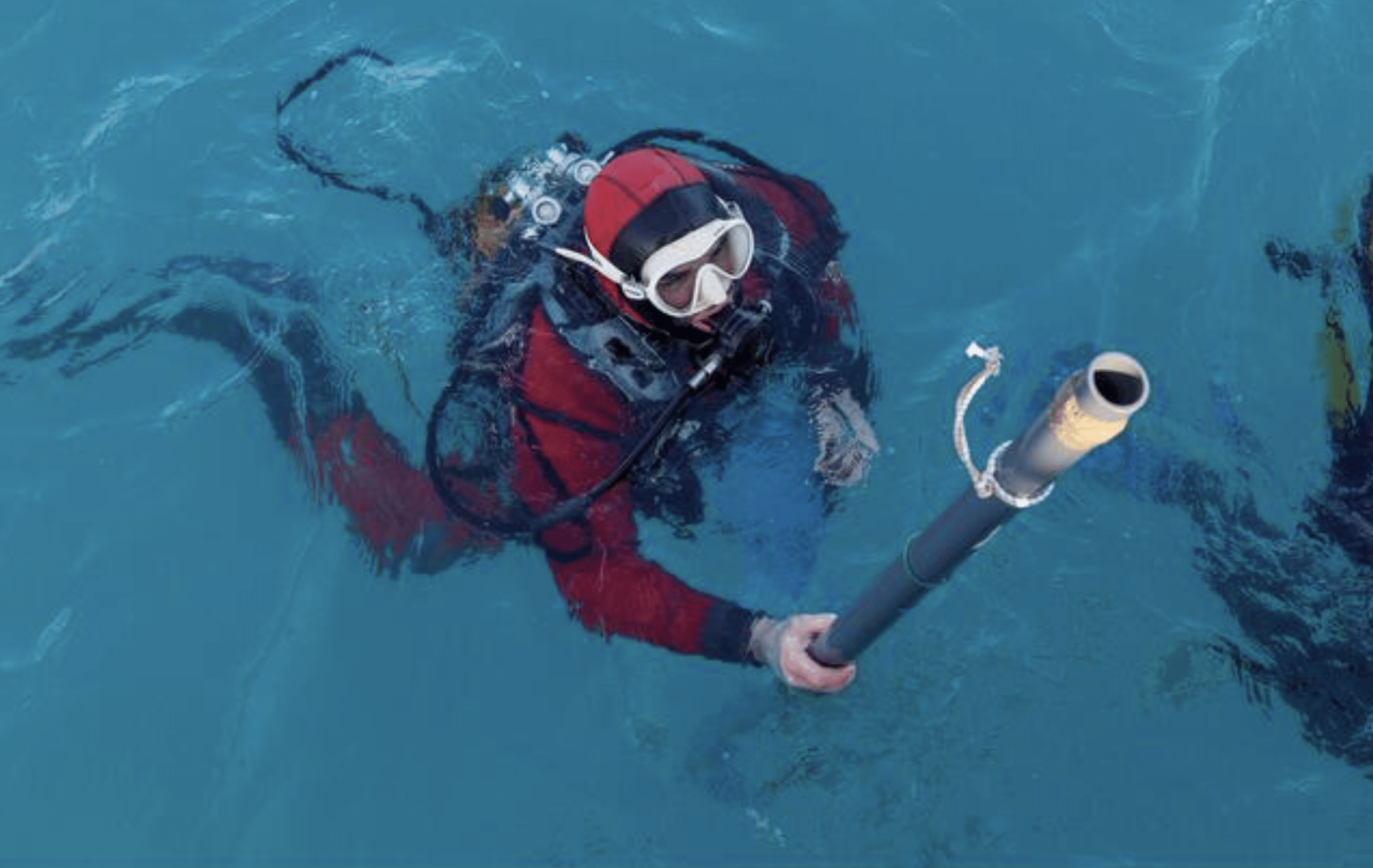 A scuba diver in a red suit and goggles is partially submerged in clear blue water, holding a long pole-like instrument upright.