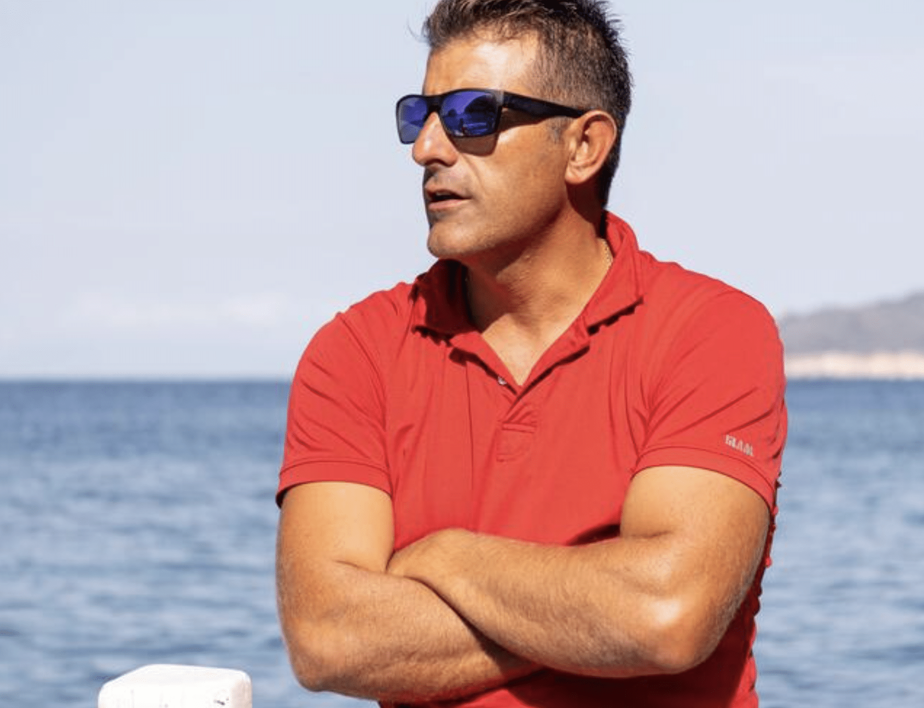 A man wearing a red polo shirt and dark sunglasses stands with arms folded in front of a calm sea, with a distant shoreline visible in the background.
