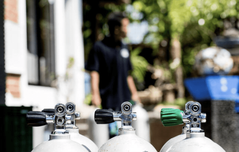 Three scuba diving air cylinders are in sharp focus in the foreground, while a person in a dark shirt stands blurred in the sunlit background amid greenery.