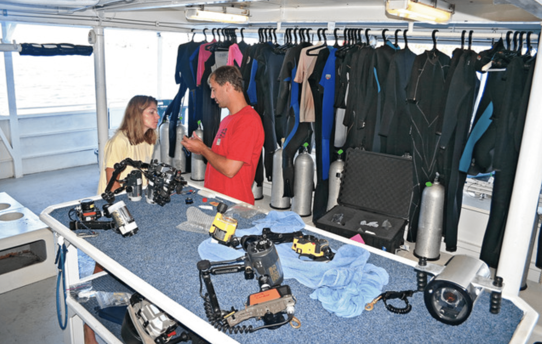 Screenshot 2026-04-06 at 232423 - N9BO℠ | Global Underwater Services Ltd Two people talk on a dive boat’s equipment table, surrounded by scuba cylinders, wetsuits, underwater cameras, and diving gear, with the sea visible outside.