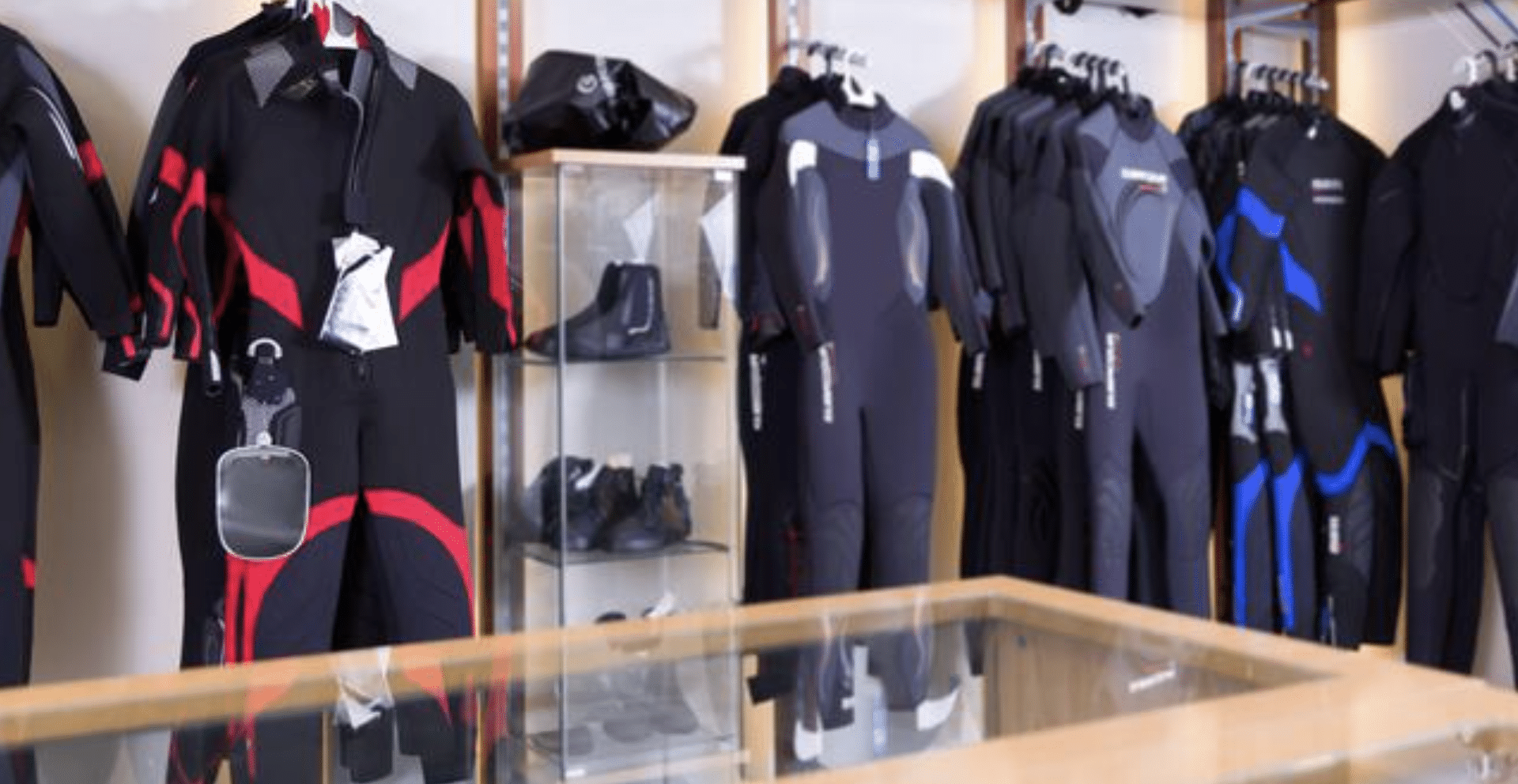 Screenshot 2026-04-06 at 232537 - N9BO℠ | Global Underwater Services Ltd Several wetsuits in black with red, blue, and grey accents hang on rails in a shop. A glass display case shows water shoes and accessories, and wetsuits are also arranged behind the case.