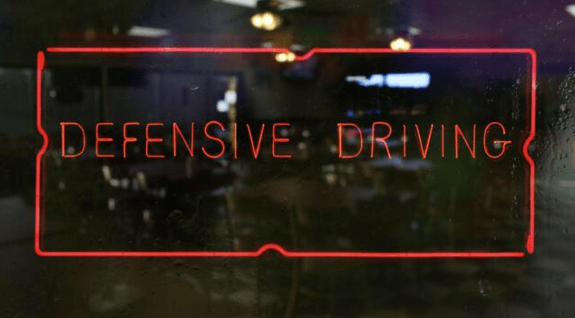 A red neon sign reads DEFENSIVE DRIVING against a dark, reflective window with a faint view of chairs and tables inside.