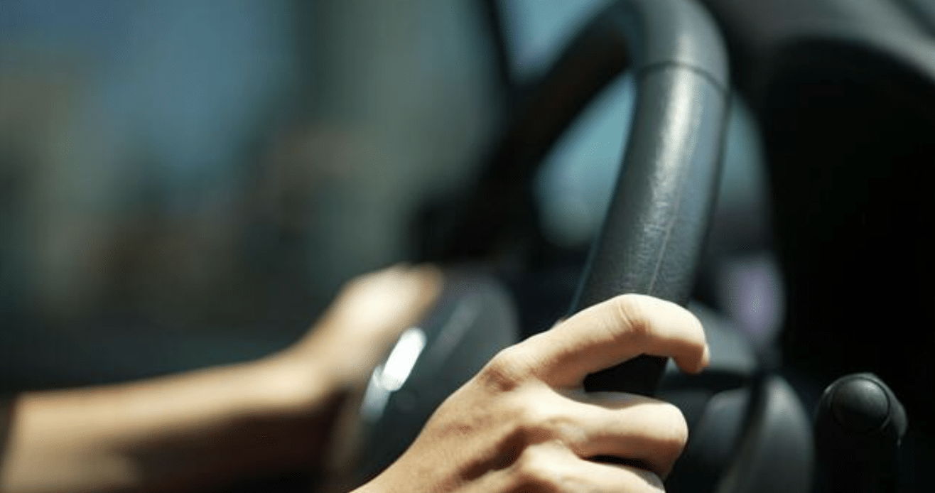 A close-up of a person’s hands gripping a car steering wheel, with sunlight illuminating their skin, suggesting they are driving during the day.