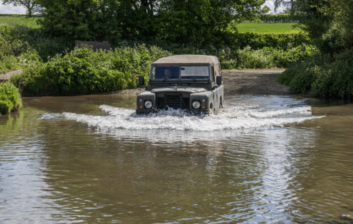 A green off-road vehicle drives through a shallow river, creating splashes, with greenery and trees in the background under a partly cloudy sky.