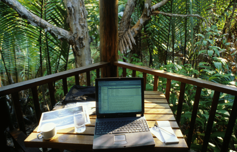 Screenshot 2026-04-07 100804 - N9BO℠ | Global Underwater Services Ltd A laptop, cup of coffee, glass of water, papers, and pen sit on a wooden table on an outdoor terrace surrounded by lush green tropical plants and trees.