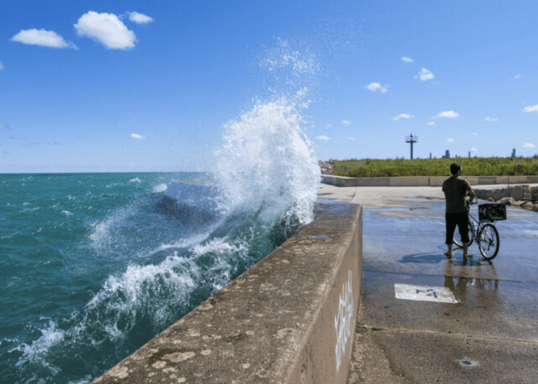 A person standing next to a bicycle watches large waves crash against a concrete pier, sending water spraying high into the air under a bright blue sky.