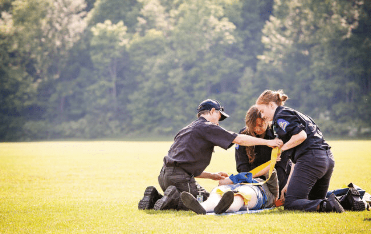 Three emergency responders kneel on grass, helping a person lying on the ground by securing their leg in a yellow splint. Trees are visible in the background.