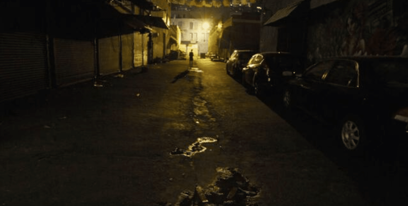 A dimly lit, empty urban alley at night with closed shop shutters on the left, parked cars on the right, and a distant figure illuminated by yellow streetlights at the far end.