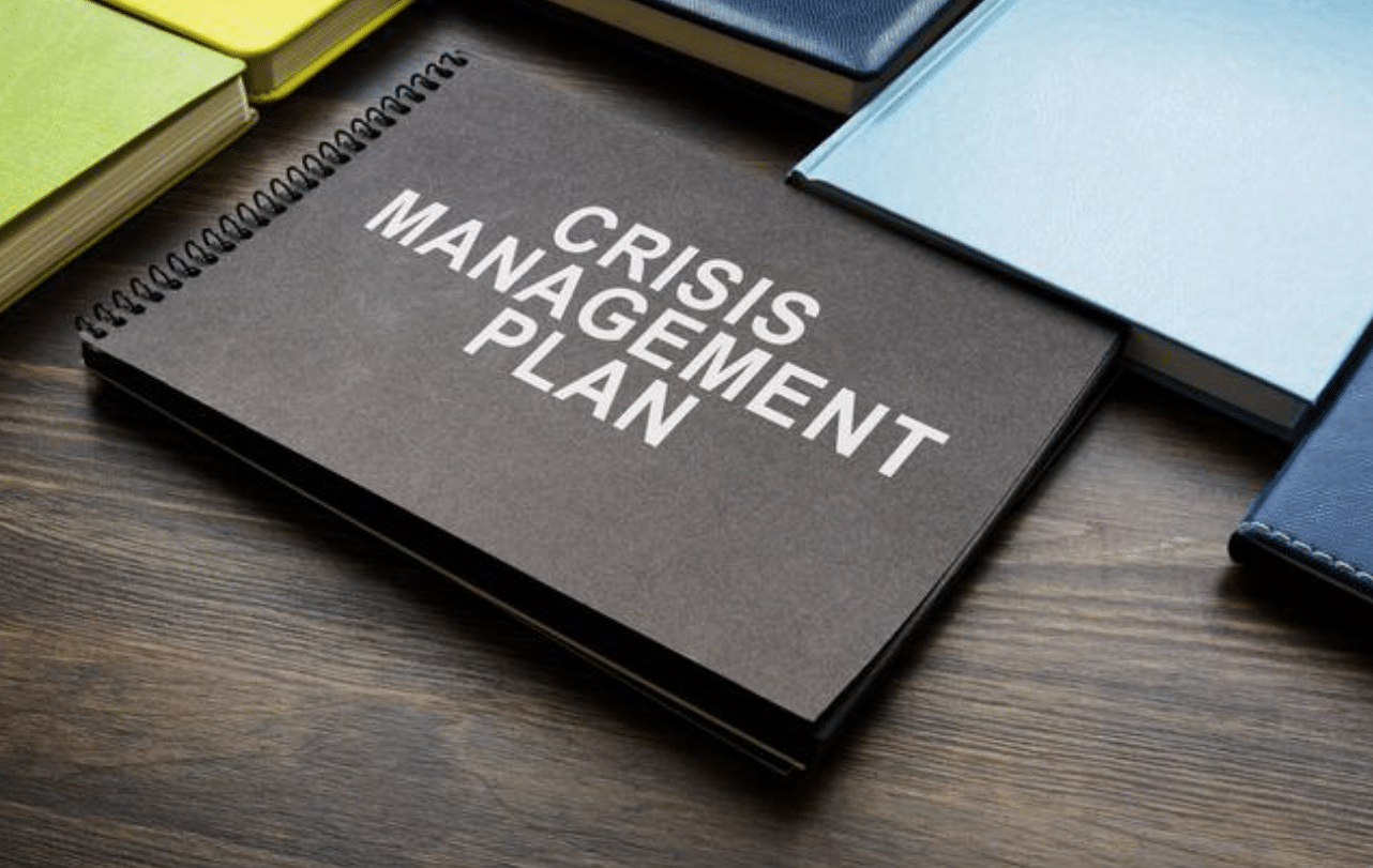 A black spiral-bound notebook labelled Crisis Management Plan sits on a wooden desk surrounded by several closed books with different coloured covers.