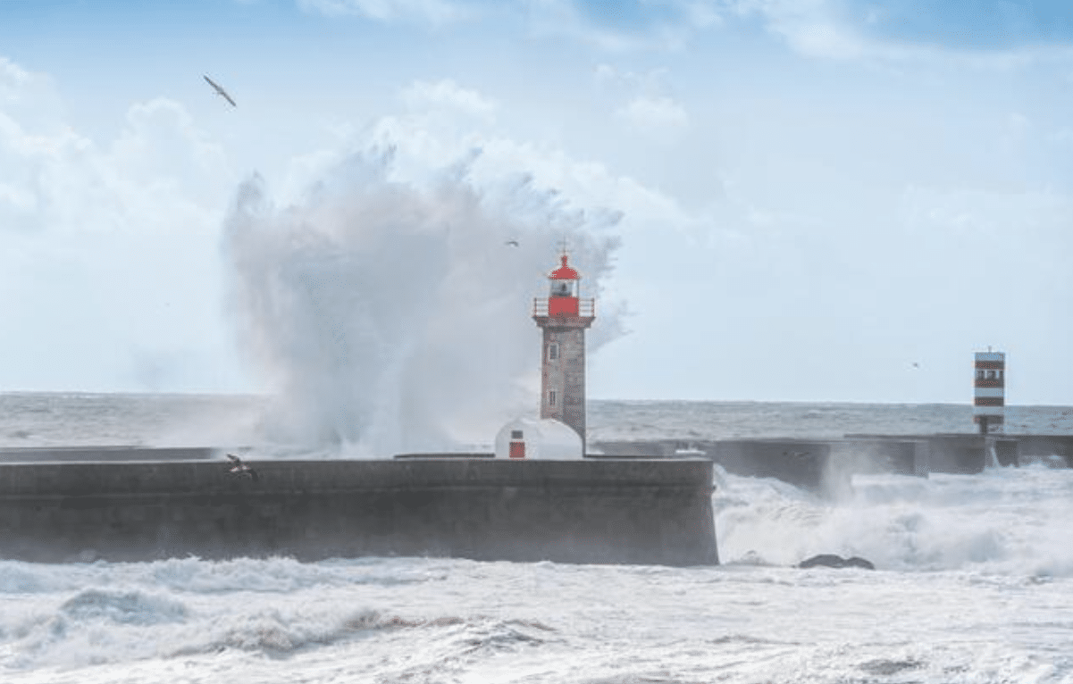 Large waves crash dramatically against a pier with two lighthouses, sending white spray high into the air, whilst seagulls fly above the rough, choppy sea under a bright, partly cloudy sky.