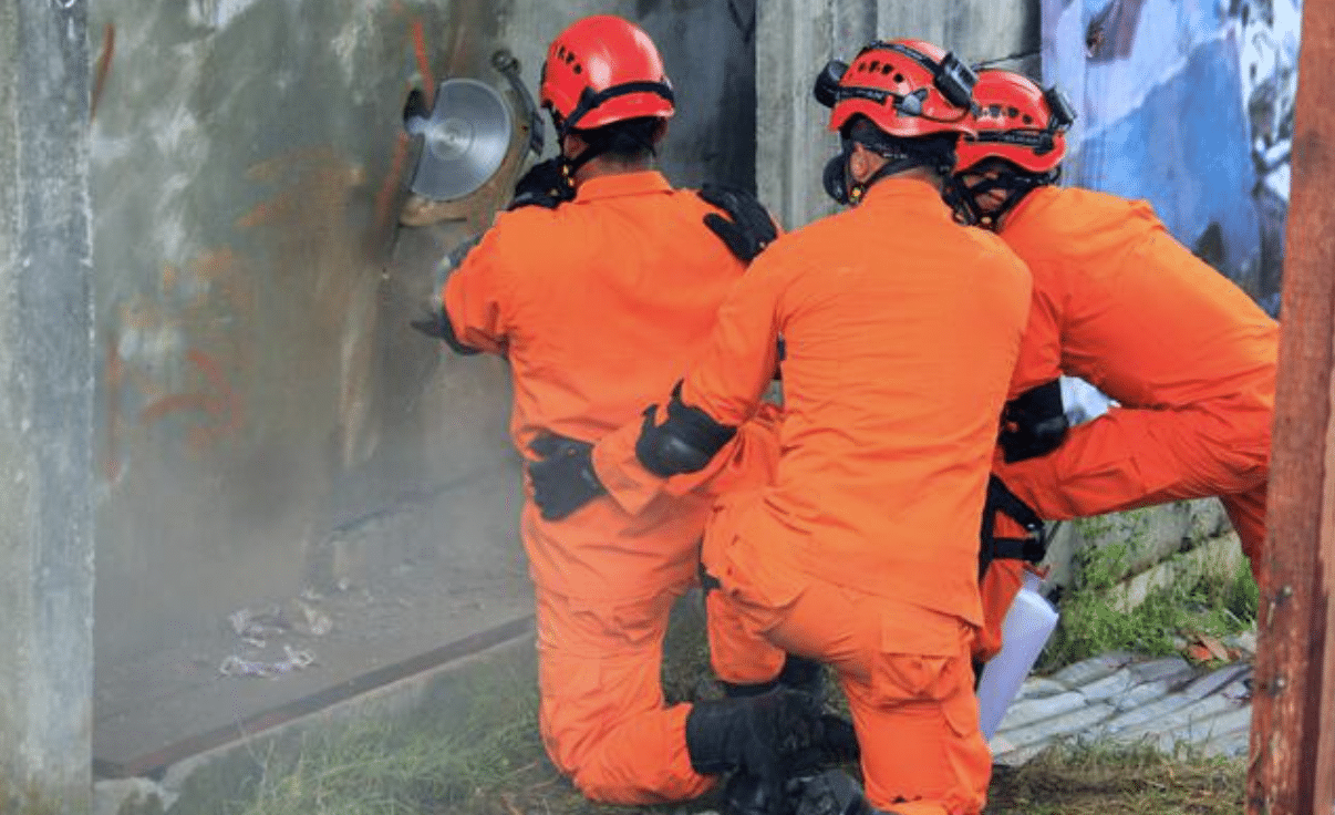 Three people in orange protective suits and helmets kneel beside a wall while one uses a circular saw to cut through concrete, with dust rising from the work.