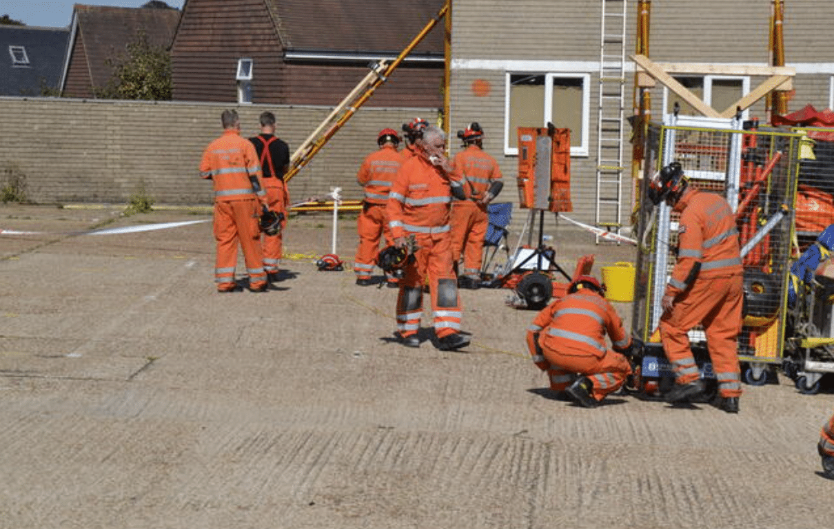 Several people wearing orange safety uniforms and helmets are gathered in an outdoor area near building materials and equipment, appearing to prepare for or participate in emergency response or rescue training.
