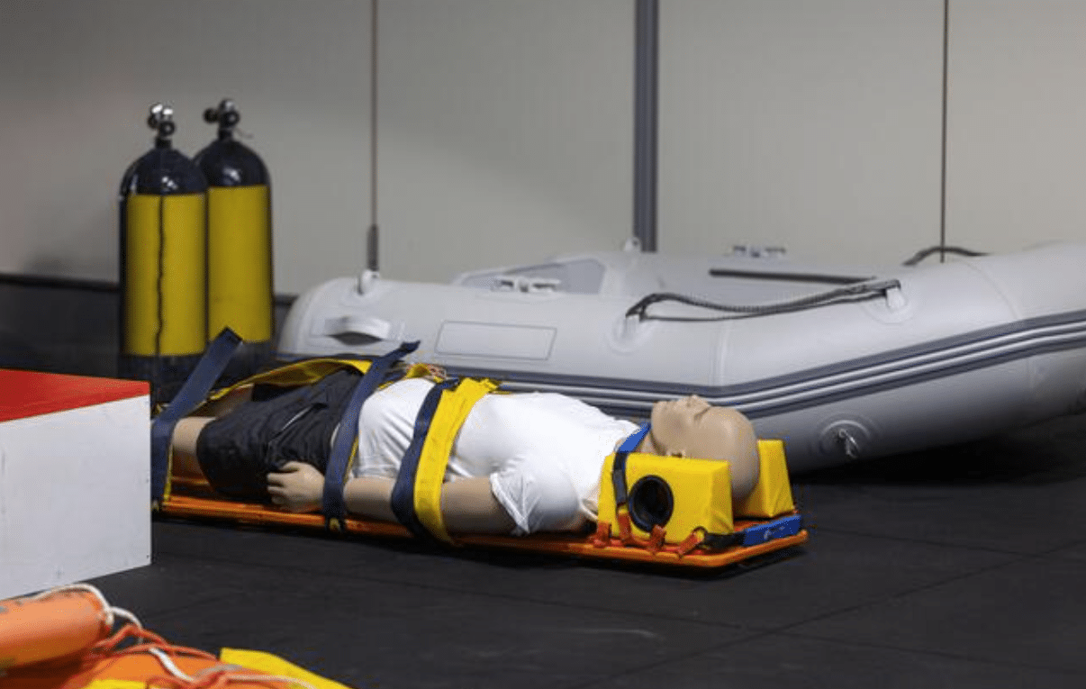 A medical training manikin strapped to a stretcher with head and neck support, lying indoors near a deflated inflatable boat and two yellow oxygen cylinders.