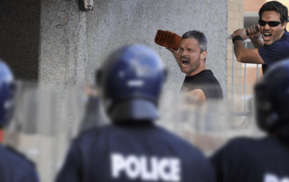 Two men shout and gesture aggressively, one holding a brick, facing a line of police officers in riot gear with shields. The police have POLICE written on their uniforms. The scene appears tense and confrontational.