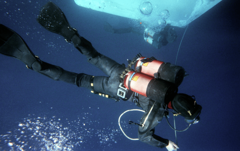 Screenshot 2026-04-07 130545 - N9BO℠ | Global Underwater Services Ltd A scuba diver in a wetsuit and fins swims underwater near a large object, holding a rope. Bubbles rise towards the surface, and another diver is visible in the background.