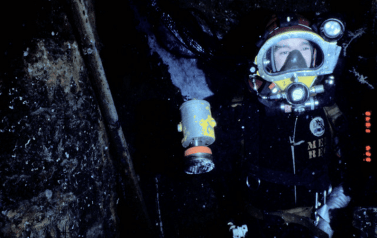 A person wearing a full diving suit and helmet holds a cylindrical object underwater, surrounded by dark, rocky surfaces and some bubbles.