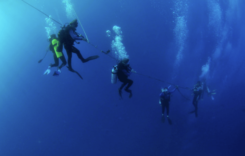 Six scuba divers descend underwater, holding onto a horizontal rope with bubbles rising to the surface against a deep blue ocean background.