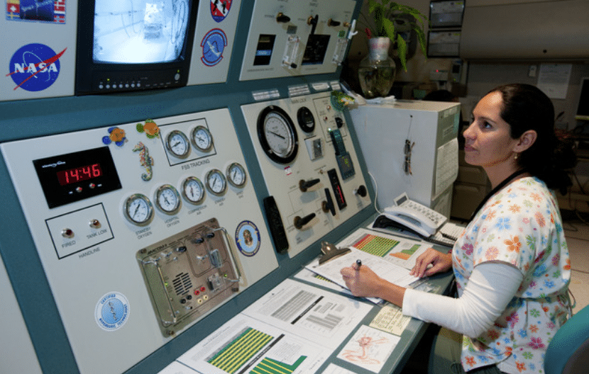 A woman sits at a control panel filled with gauges, dials, and screens, taking notes on papers. A NASA sticker is visible, and a monitor shows a room, suggesting a scientific or technical environment.