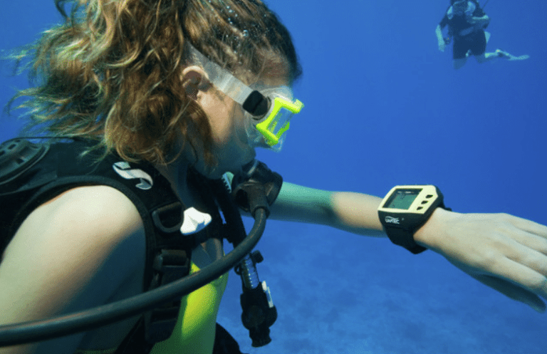 A scuba diver with long, curly hair and yellow goggles checks a dive computer on her wrist underwater. Another diver is visible in the blue water background.