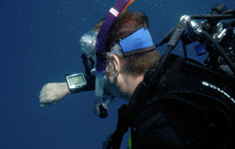 A scuba diver underwater wearing a snorkel and looking at a dive computer on their wrist, surrounded by deep blue water.