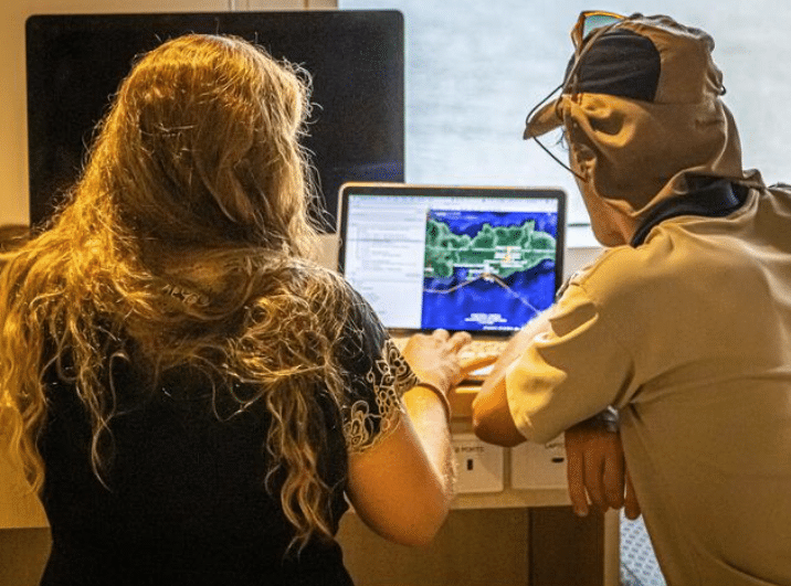 Two people sit side by side at a desk, looking at a laptop displaying a map or simulation on the screen. One person wears a light brown hat with ear flaps; both are focused on the computer.