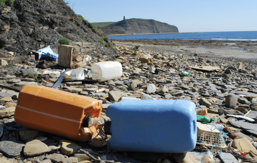 Screenshot 2026-04-07 145409 - N9BO℠ | Global Underwater Services Ltd Plastic containers and various debris scattered across a rocky beach with a cliff and the sea in the background under a clear blue sky.