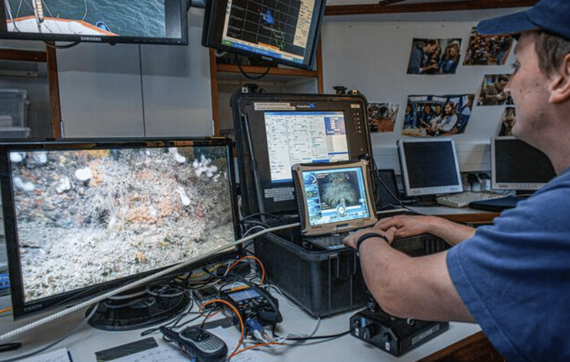 Screenshot 2026-04-07 160641 - N9BO℠ | Global Underwater Services Ltd A person operates underwater exploration equipment, monitoring seabed footage on multiple screens in a control room filled with computers, cables, and oceanographic instruments.