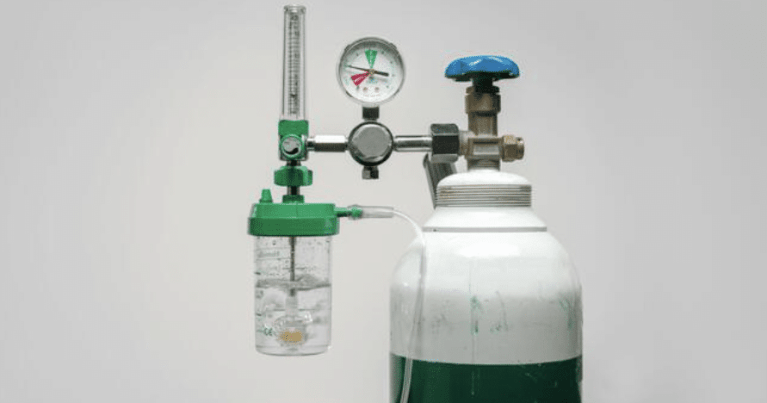 A medical oxygen cylinder with a pressure gauge, flow meter, and humidifier bottle attached to the regulator, set against a plain light grey background.