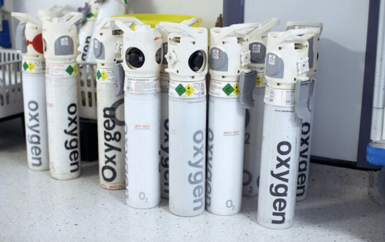 Screenshot 2026-04-07 170357 - N9BO℠ | Global Underwater Services Ltd Several white oxygen cylinders with labelled oxygen stand upright on a floor. They have gauges and valves on top, and warning labels are visible on the tanks. The background includes shelves and other equipment.