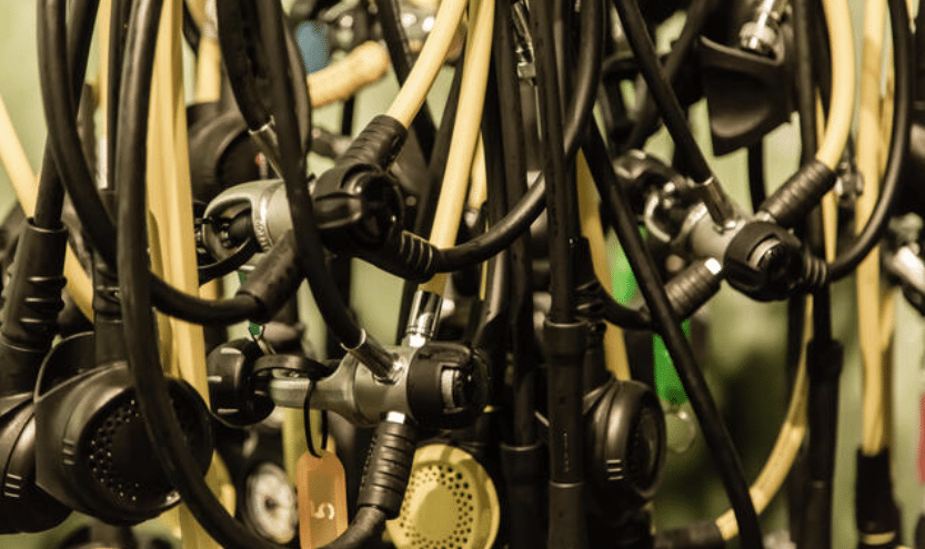 Screenshot 2026-04-07 171008 - N9BO℠ | Global Underwater Services Ltd A close-up of several scuba diving regulators and hoses hanging together, showing mouthpieces, gauges, and tags, with the equipment appearing tangled and densely packed.