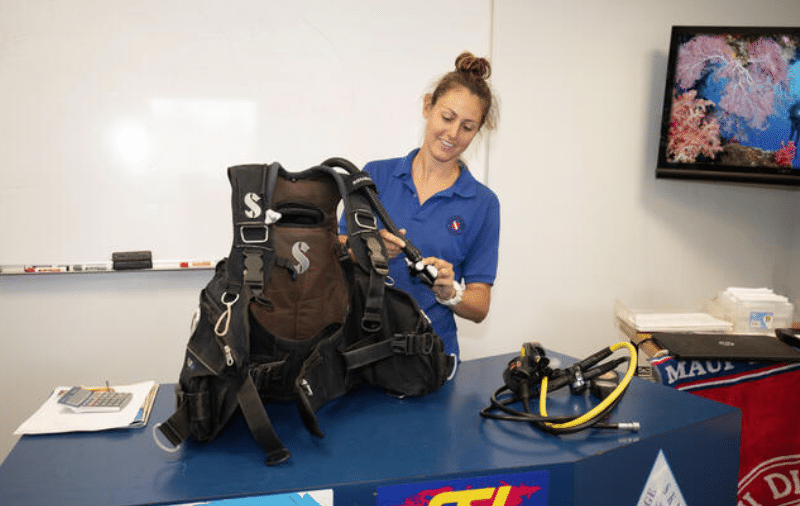 Screenshot 2026-04-07 171023 - N9BO℠ | Global Underwater Services Ltd A woman in a blue shirt stands behind a counter, examining scuba diving equipment, with a TV displaying coral reefs and a whiteboard in the background.