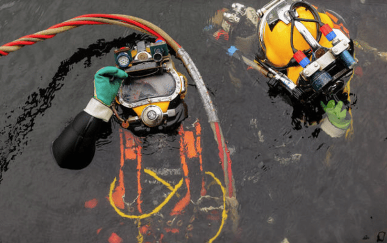 Two commercial divers in yellow helmets and protective gear float in dark water, holding cables and hoses. One diver is partially submerged, holding his helmet, while the other has both hands visible above the water.