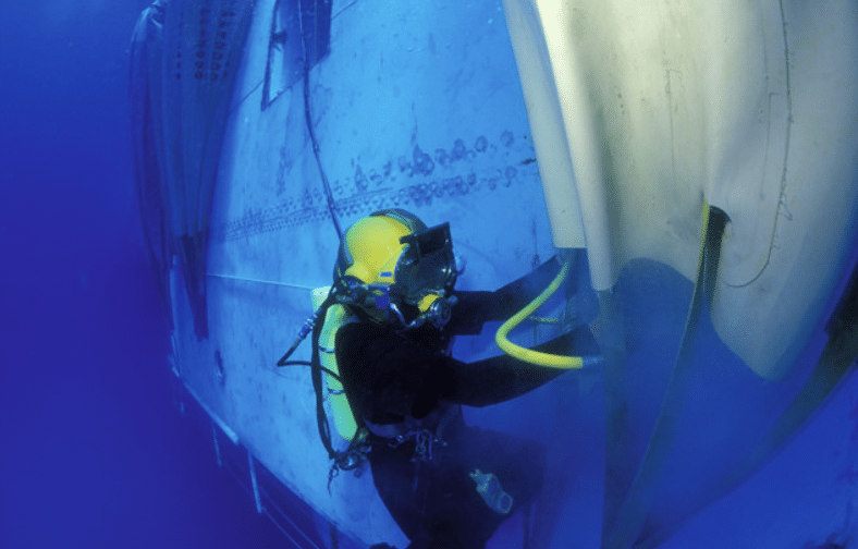 Screenshot 2026-04-07 171649 - N9BO℠ | Global Underwater Services Ltd A scuba diver in a yellow helmet and black wetsuit works underwater, inspecting or repairing the side of a large submerged structure, possibly a ship or submarine, whilst holding a yellow hose.