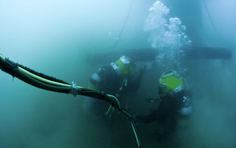 Screenshot 2026-04-07 171701 - N9BO℠ | Global Underwater Services Ltd Two deep-sea divers in yellow helmets work underwater near thick cables and a large pipe, with air bubbles rising to the surface in the murky, blue-green water.